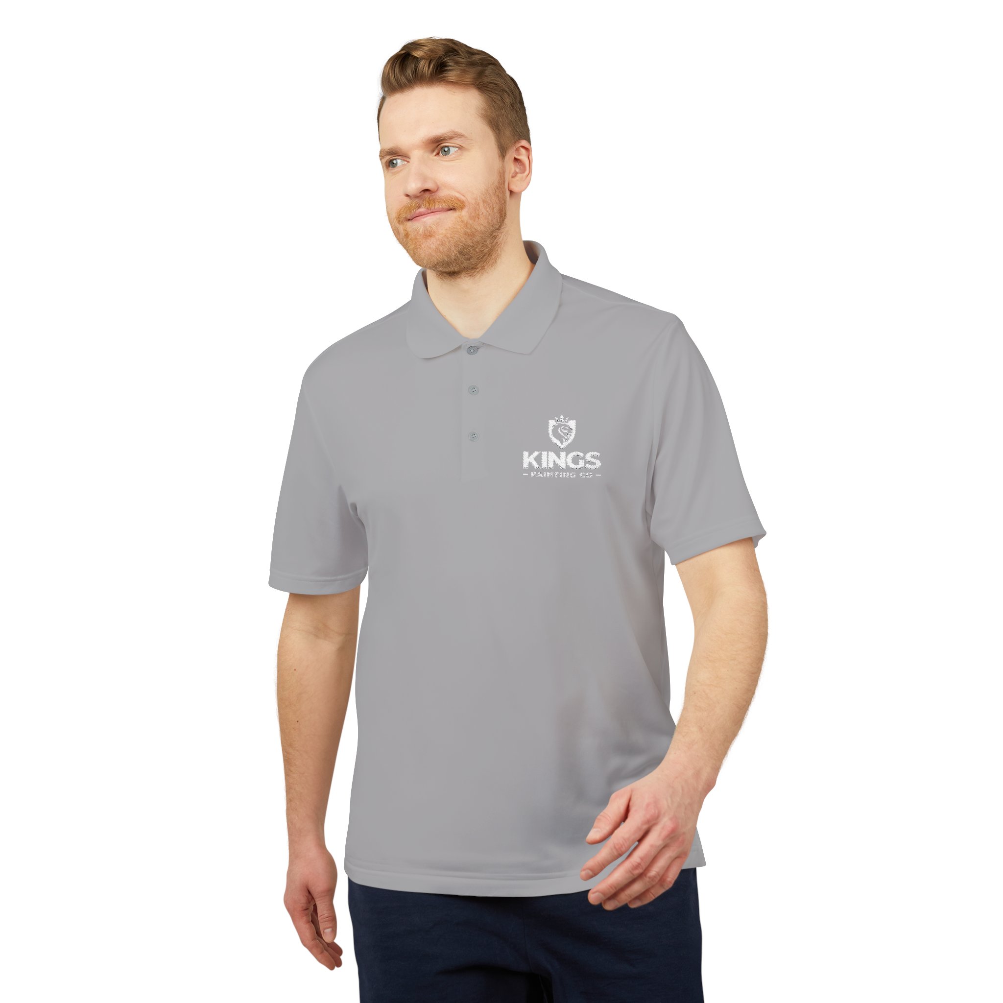 A man with light brown hair and a beard wears the Kings Painting Co. Performance Polo by Under Armour®, featuring a white KINGS crown logo on the left chest, as he stands looking to the side against a white background.